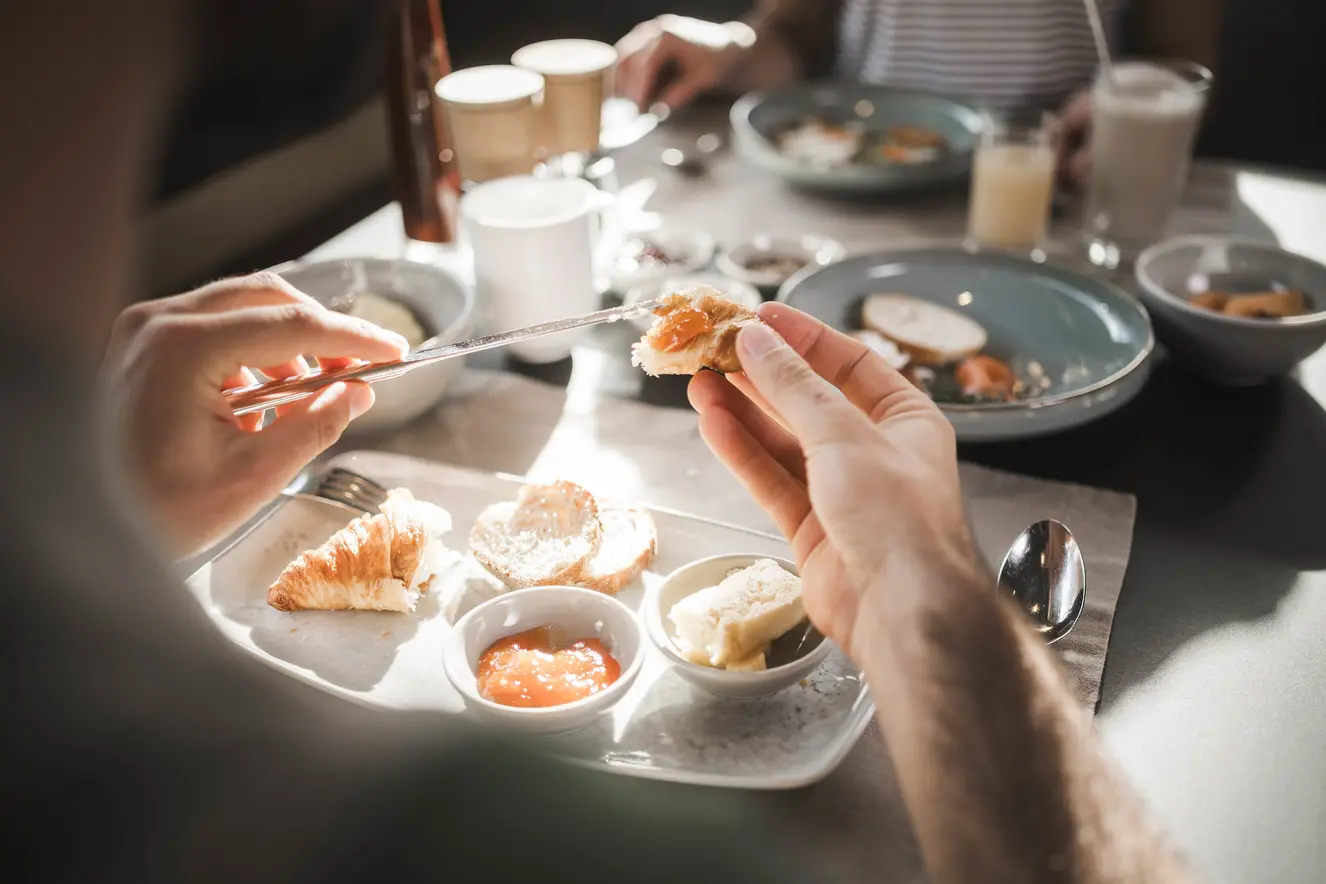 Eine Person bestreicht ein Croissant mit Butter an einem Frühstückstisch mit Marmelade, Kaffee und Saft.
