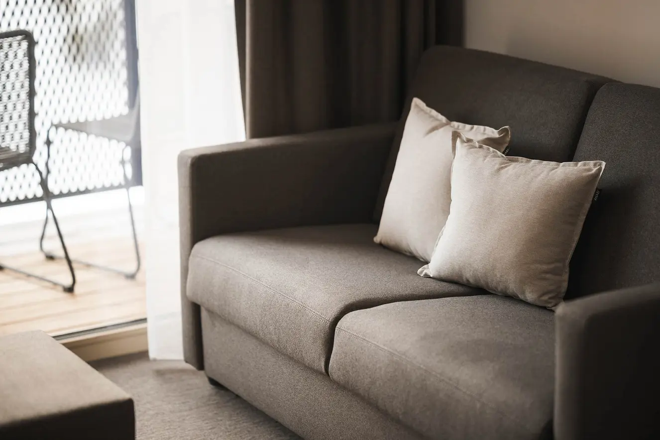 Ein graues Sofa mit zwei beigen Kissen in der Nähe eines Fensters und draußen ein Balkon mit einem schwarzen Stuhl.