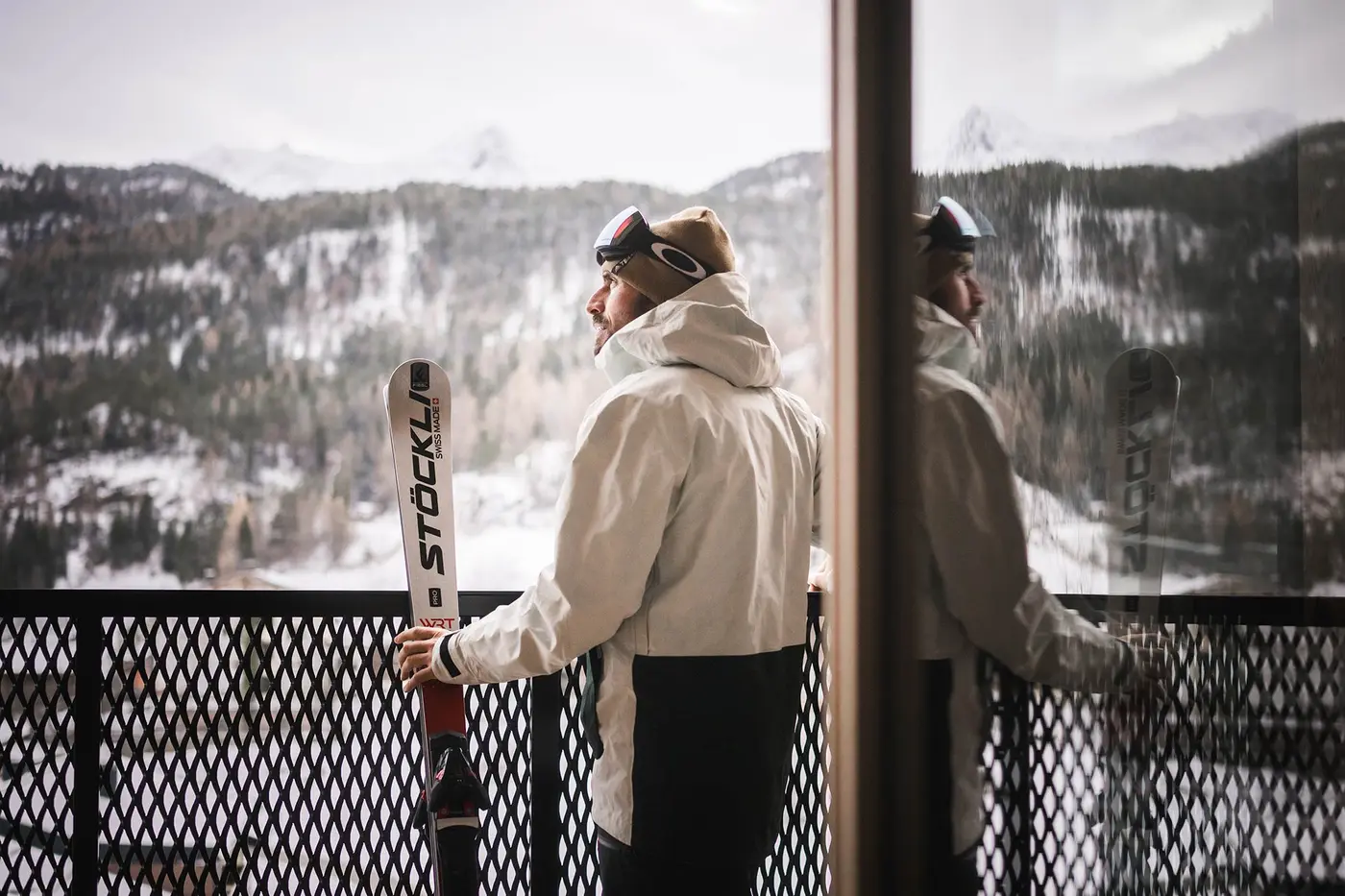 Eine Person in Skiausrüstung steht mit Skiern in der Hand auf einem Balkon und blickt auf schneebedeckte Berge, deren Spiegelbild sich im Glas spiegelt.