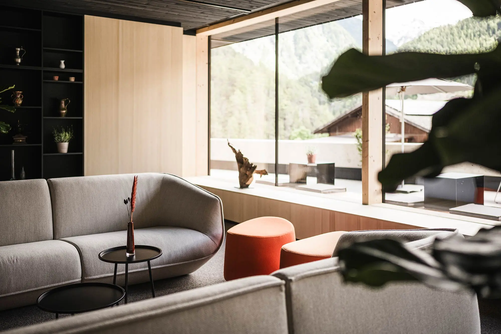 Modernes Wohnzimmer mit hellgrauen Sofas, runden Tischen, orangefarbenen Ottomanen, großem Fenster und Blick auf die Berge.