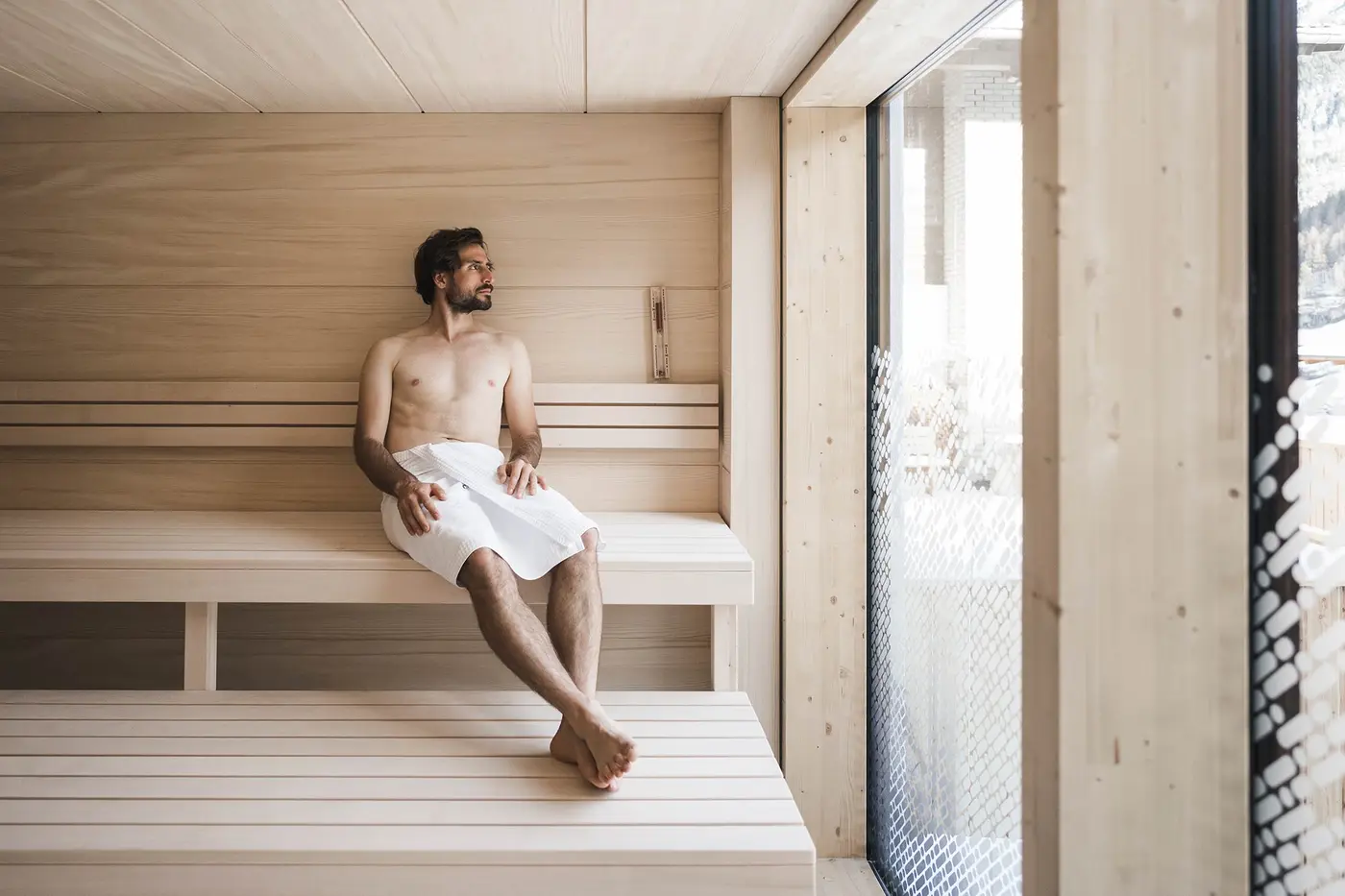Ein Mann mit Handtuch sitzt entspannt auf einer Holzbank in einer modernen Sauna und blickt aus einem großen Fenster.