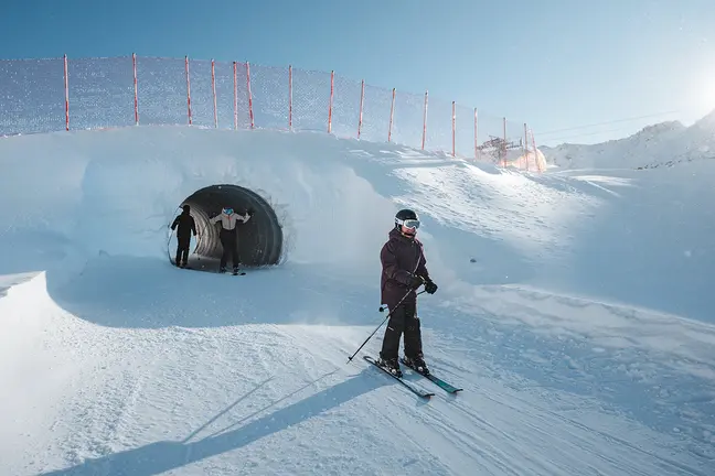 Drei Personen fahren Ski auf Schnee; zwei verlassen einen Tunnel, während eine im Vordergrund unter einem klaren blauen Himmel Ski fährt.