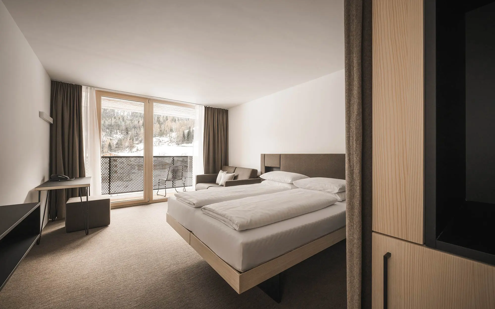 Minimalistisches Hotelzimmer mit Doppelbett, Schreibtisch und großem Fenster mit Blick auf eine verschneite Waldlandschaft.