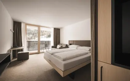 Minimalistisches Hotelzimmer mit Doppelbett, Schreibtisch und großem Fenster mit Blick auf eine verschneite Waldlandschaft.