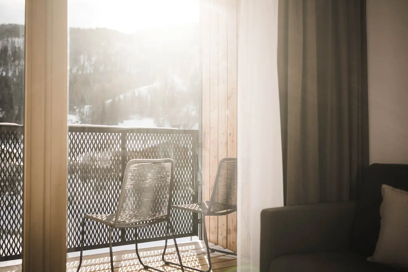 Zwei Metallstühle auf einem sonnendurchfluteten Balkon mit Blick auf eine verschneite Berglandschaft.