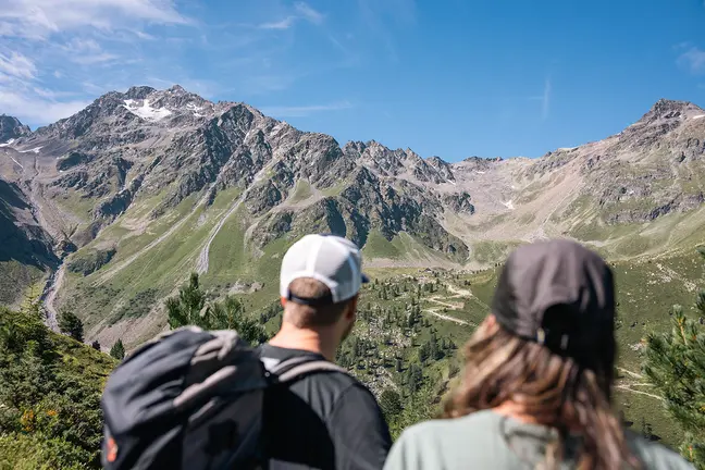 Zwei Wanderer mit Rucksäcken blicken auf eine malerische Berglandschaft unter einem klaren blauen Himmel.