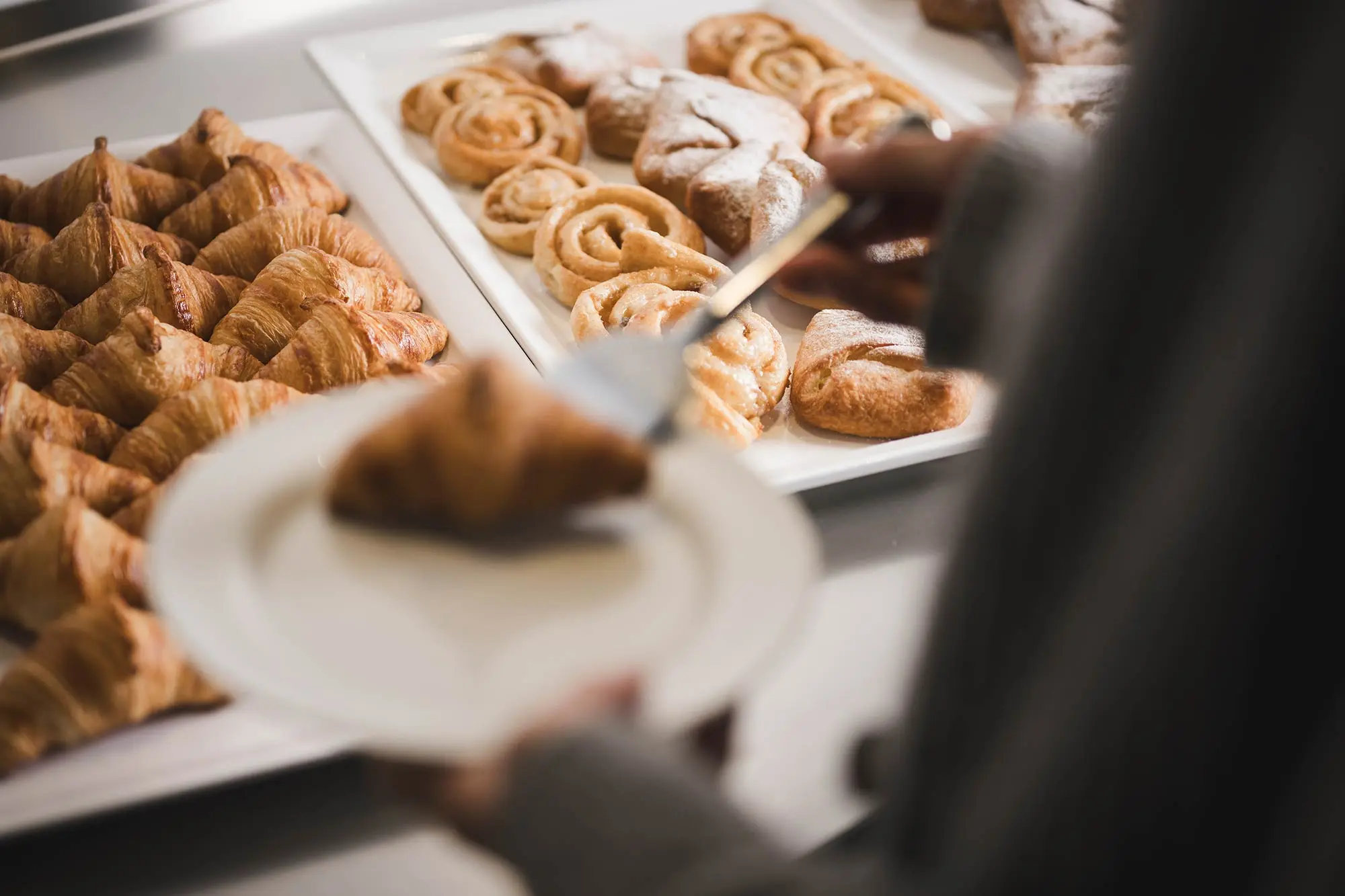 Eine Person serviert von einem Tisch mit verschiedenen Gebäckstücken ein Croissant auf einem Teller.