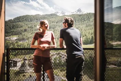 Zwei Personen unterhalten sich an einem sonnigen Tag auf einem Balkon mit malerischen Bergen und einem Wald im Hintergrund.