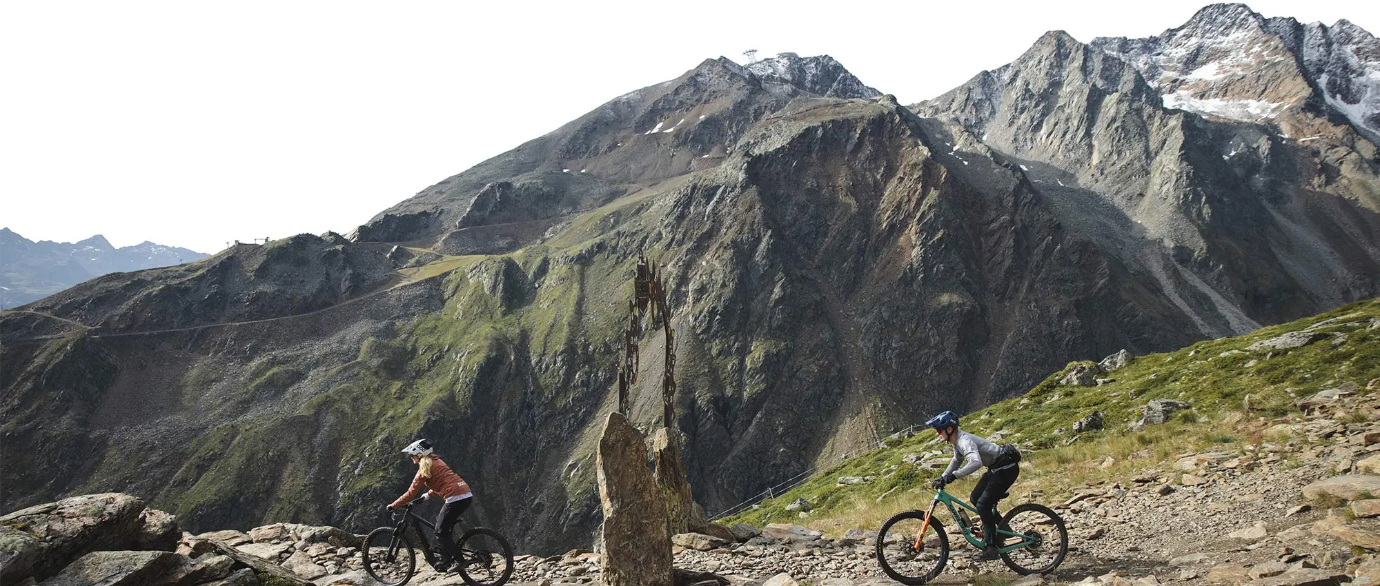 Zwei Mountainbiker fahren auf einem steinigen Pfad mit hohen, schroffen Bergen im Hintergrund.