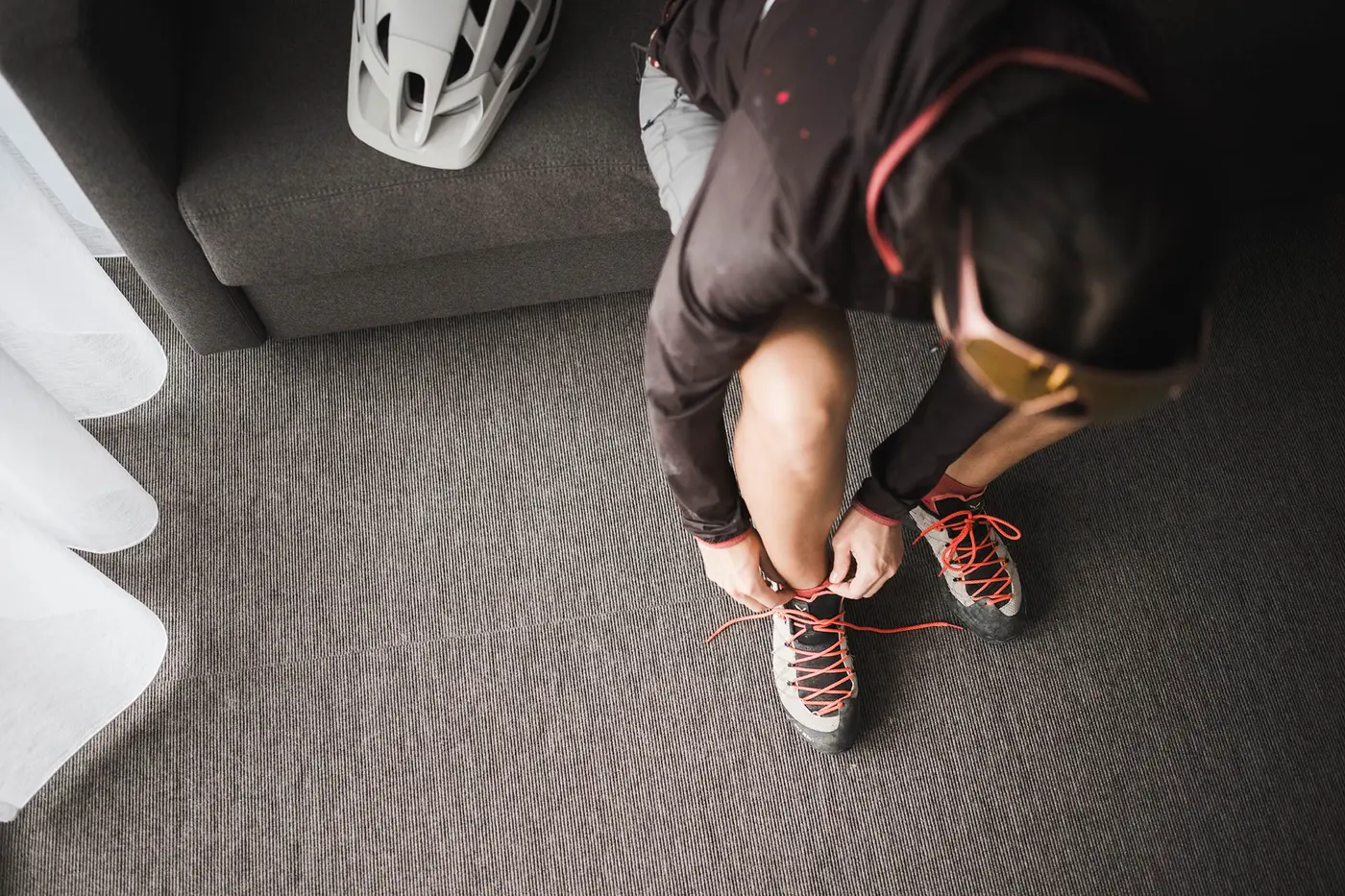 Eine Person sitzt auf einem Sofa und bindet die Schnürsenkel ihrer Wanderschuhe. In der Nähe liegt auf dem Sofa ein Helm.